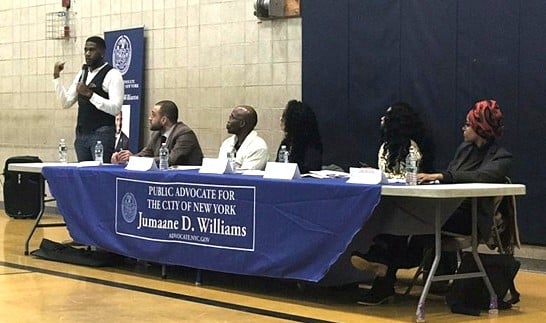 A man speaks at a podium next to a panel of people seated at a table. The table features a blue banner for New York Public Advocate Jumaane D. Williams during a bail reform event co-sponsored by Katal.