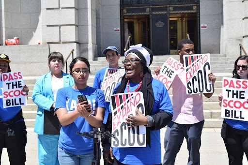 A group of advocates stands on government building steps, holding signs that say “Pass the SCOC Bill” and “Expand SCOC.” One speaker in a blue shirt and headwrap addresses the crowd while others stand in solidarity behind her.