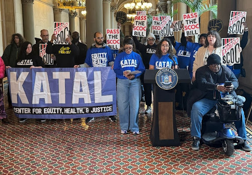 A group of advocates from the Katal Center for Equity, Health, and Justice hold a press conference indoors, standing behind a podium with the New York State seal. They wear matching blue KATAL shirts and hold a large purple banner reading “KATAL Center for Equity, Health, & Justice,” while others in the background raise signs reading “PASS THE SCOC BILL.”