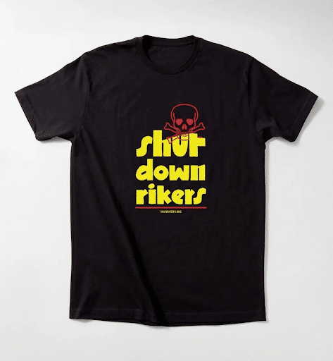 Black t-shirt with bold yellow and red text reading “shut down rikers” under a red skull and crossbones. The shirt is laid flat on a white surface and appears to be part of a protest or advocacy campaign.